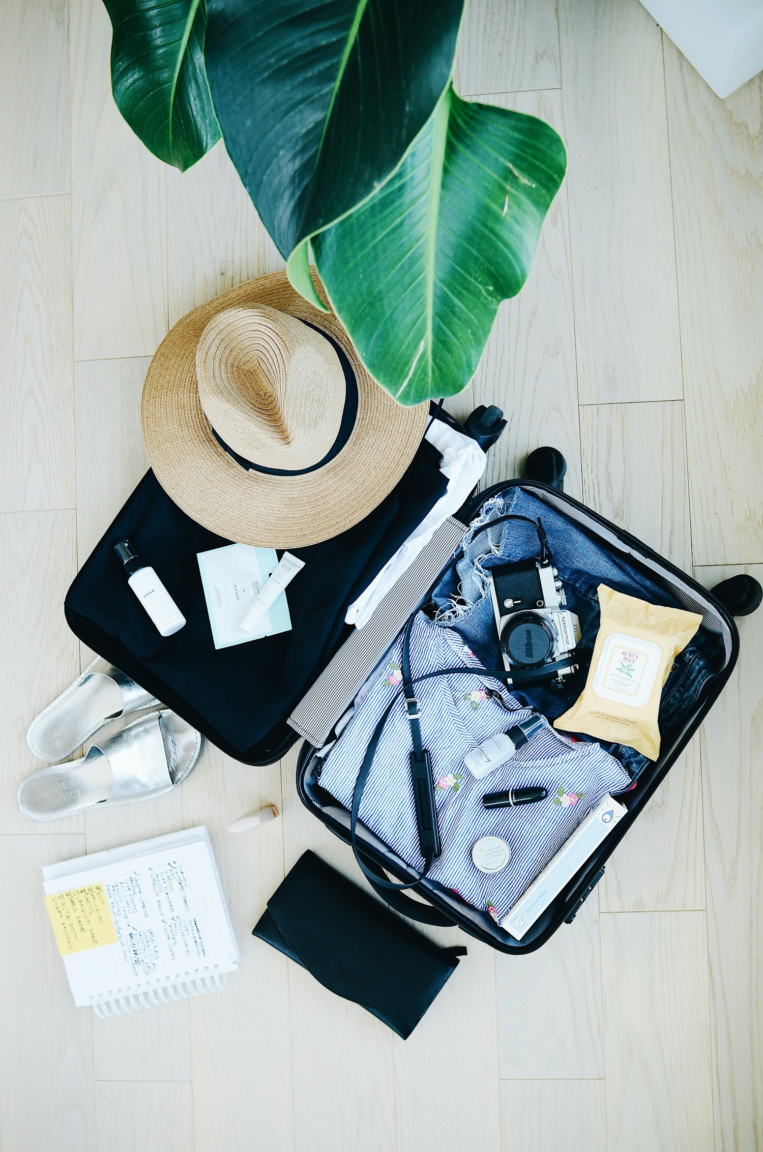 Open suitcase mid-pack with a sun hat, camera, sandals and a handwritten packing list