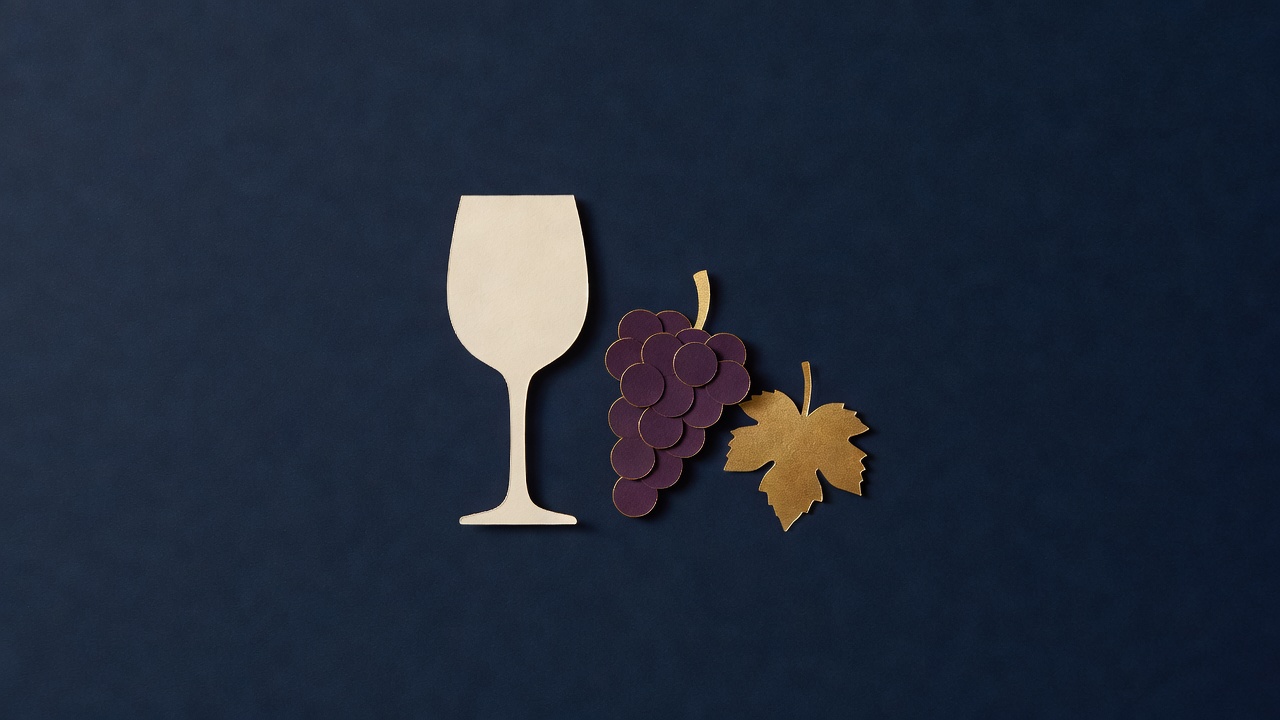 Paper-cut still life of an empty paper wine glass, a plum paper grape cluster and a gold vine leaf on navy