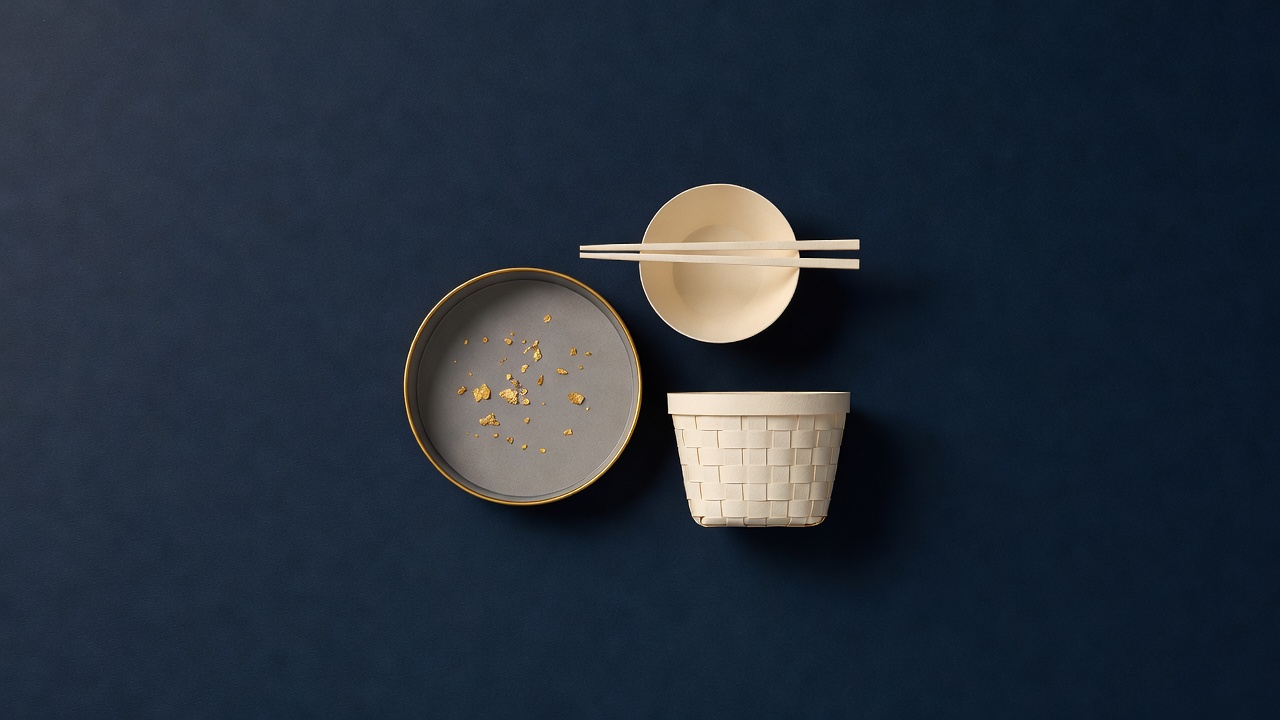 Paper-cut still life of a gold pan with gold flakes, a tea bowl with chopsticks and a small woven basket on navy